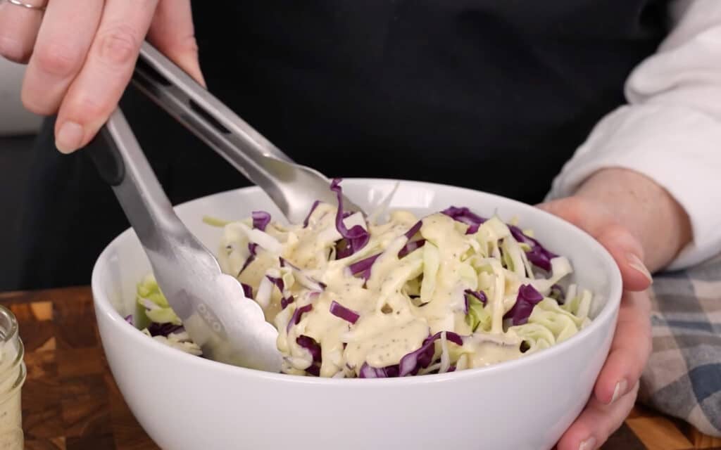A horizontally aligned action shot, showing Mary tossing shredded cabbage with the coleslaw dressing with a pair of tongs.