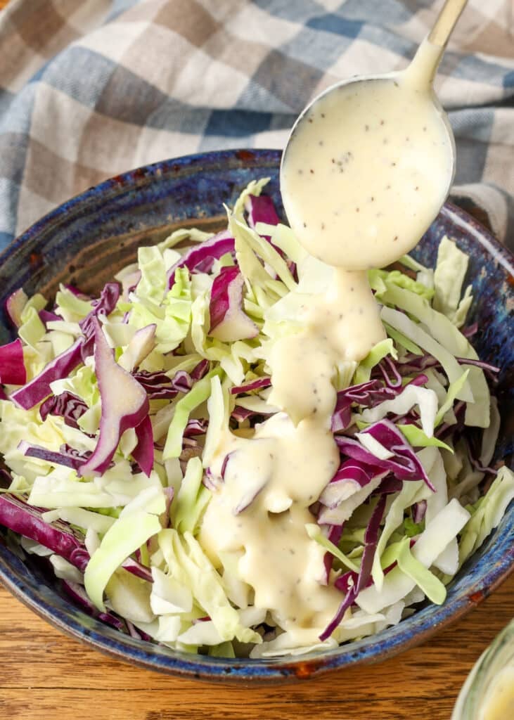 Dressing the coleslaw with a metal spoon, drizzling it over freshly shredded cabbage.