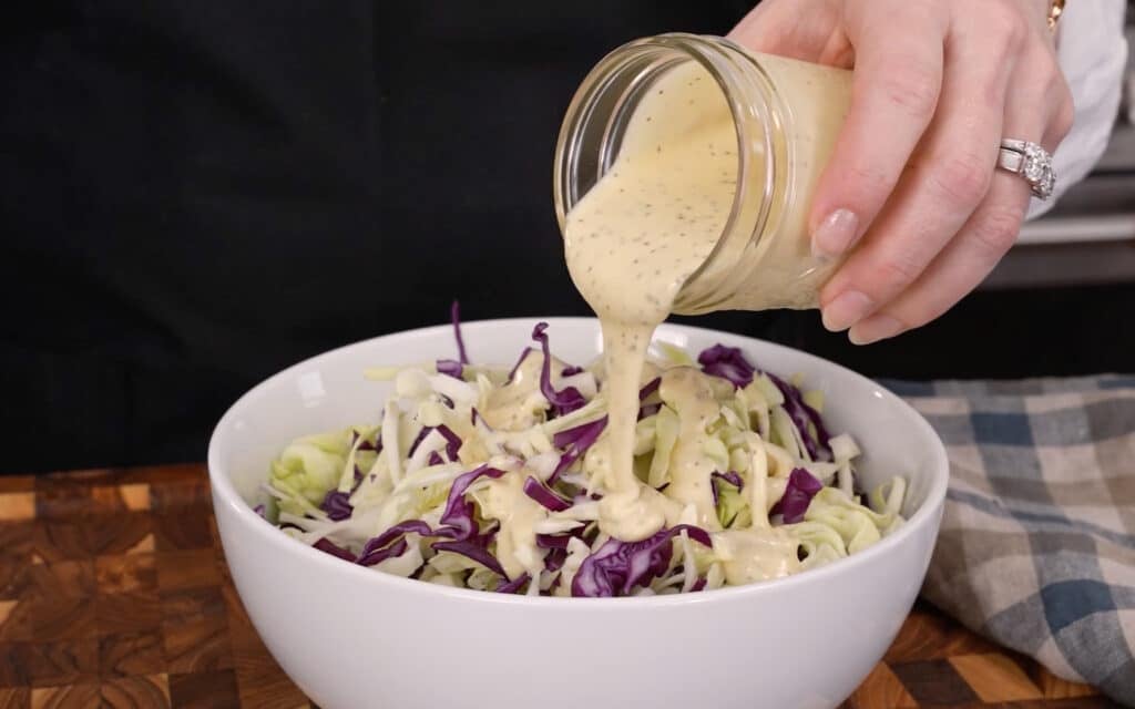 A horizontally aligned action shot showing Mary pouring the dressing from a small jar over the cabbage.