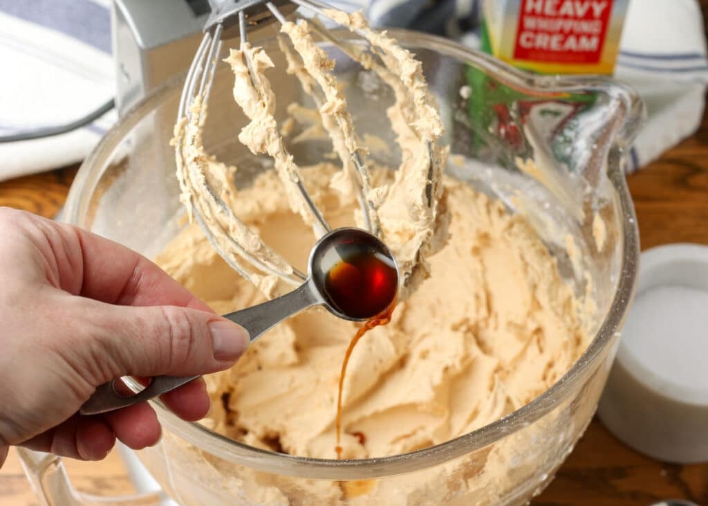 A close up of the measuring spoon drizzling the vanilla extract into a stand mixer full of soon-to-be frosting.