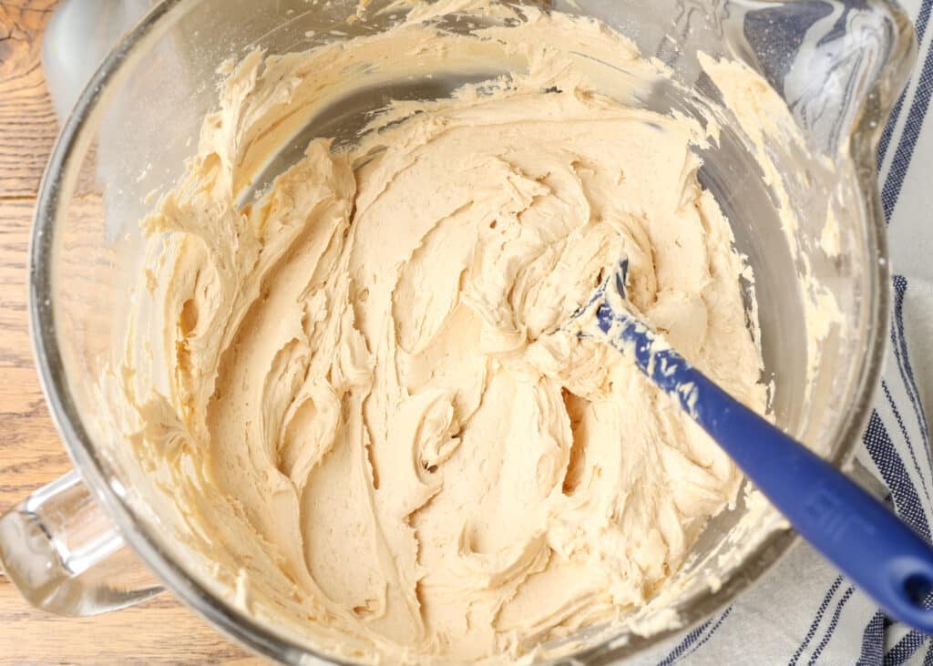 A blue spatula in a clear mixing bowl full of light brown frosting.