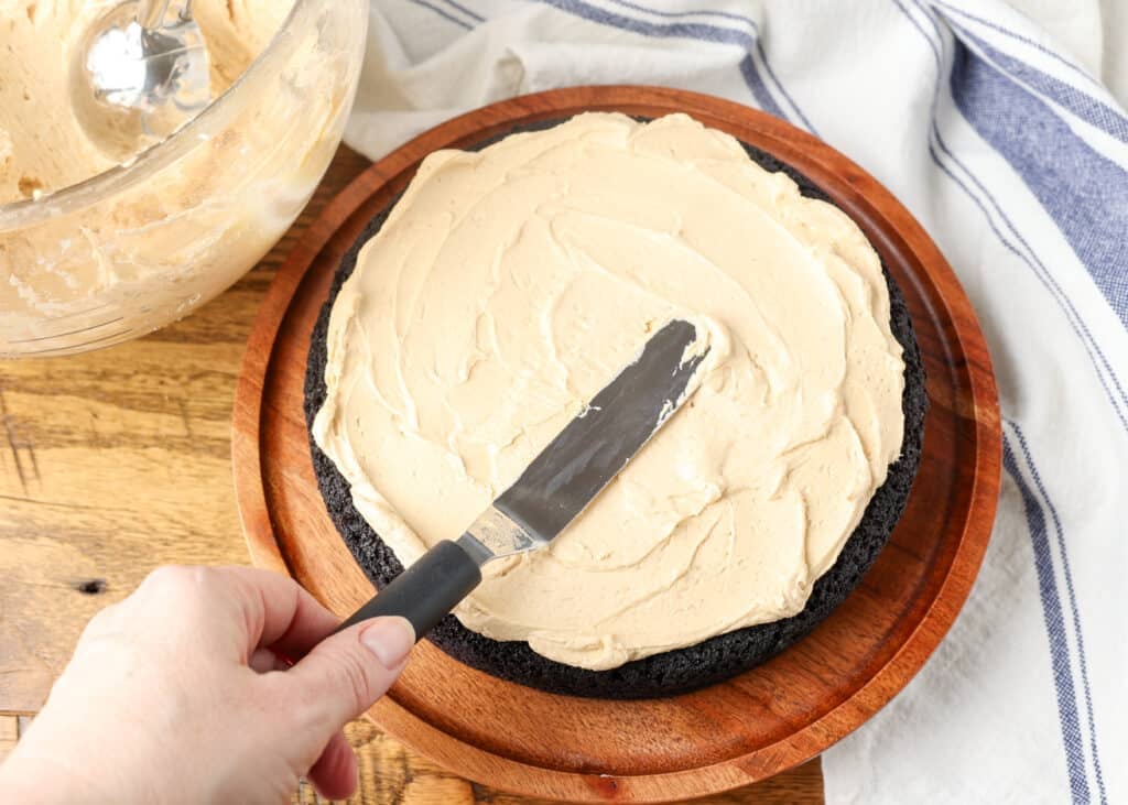 An action shot showing a hand holding the spatula, spreading the frosting over the top of a layer of round cake.