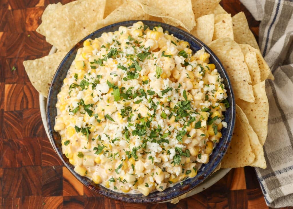 elote dip in blue bowl surrounded by chips on wooden table