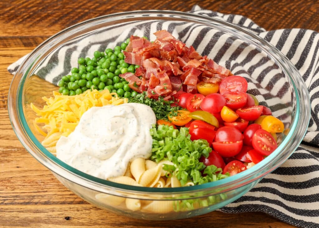 pasta salad ingredients with ranch dressing, bacon, tomatoes, green onions, peas, tomatoes, and cheese in clear mixing bowl