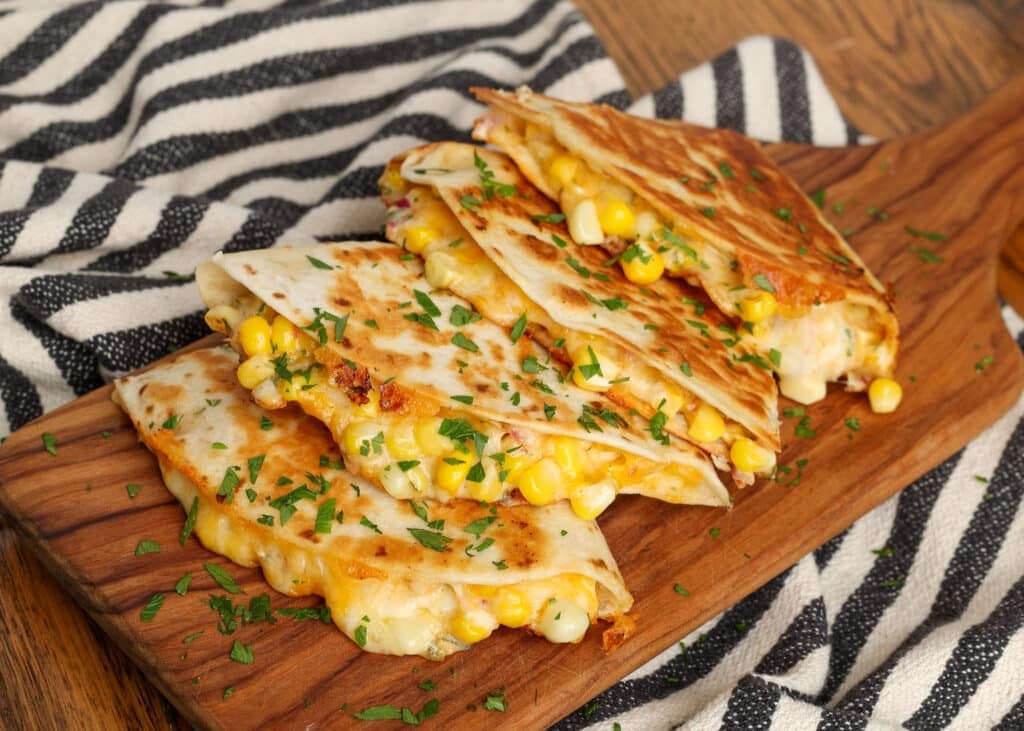 horizontal photo of elote quesadillas on wooden board with black and white striped towel